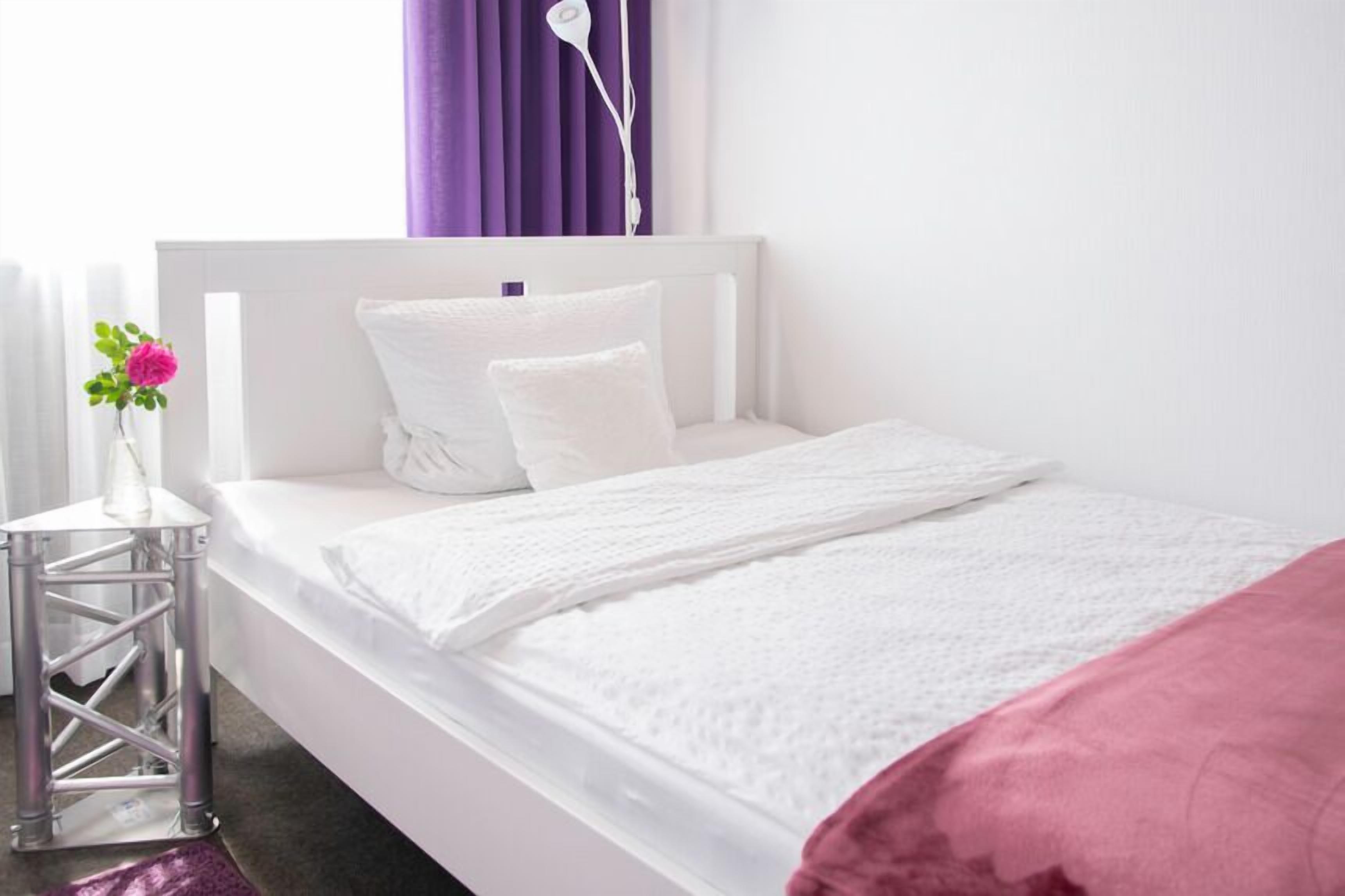Un letto bianco con una coperta rosa, una testata bianca e una tenda viola.