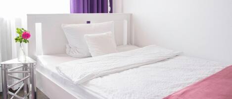 A white bed with a pink blanket, a white headboard, and a purple curtain.