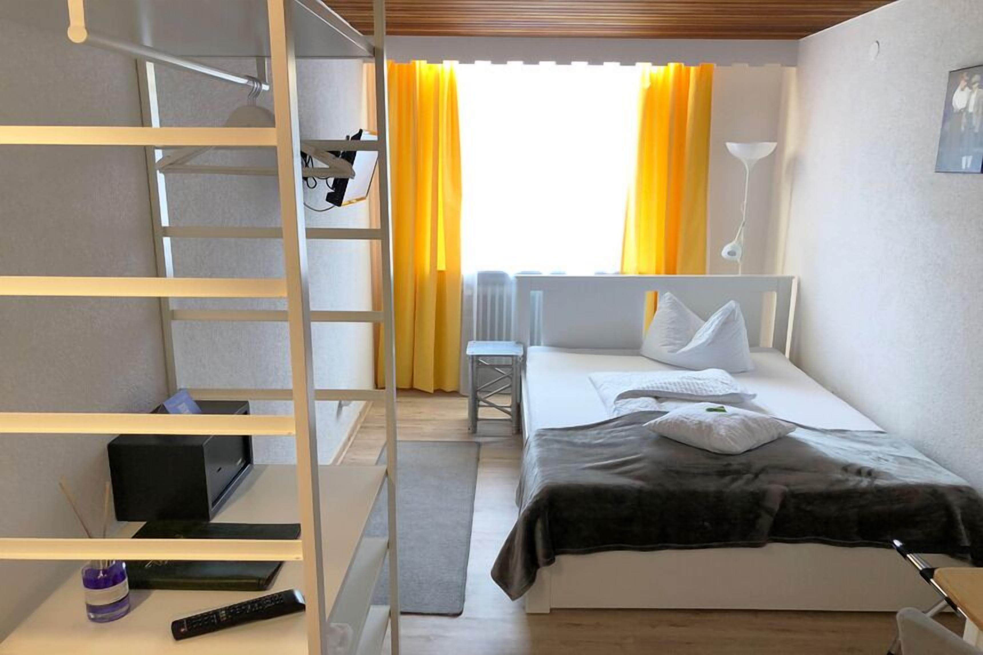 Une chambre d’hôtel comprenant un lit, un bureau, une chaise, une étagère et une fenêtre avec des rideaux.