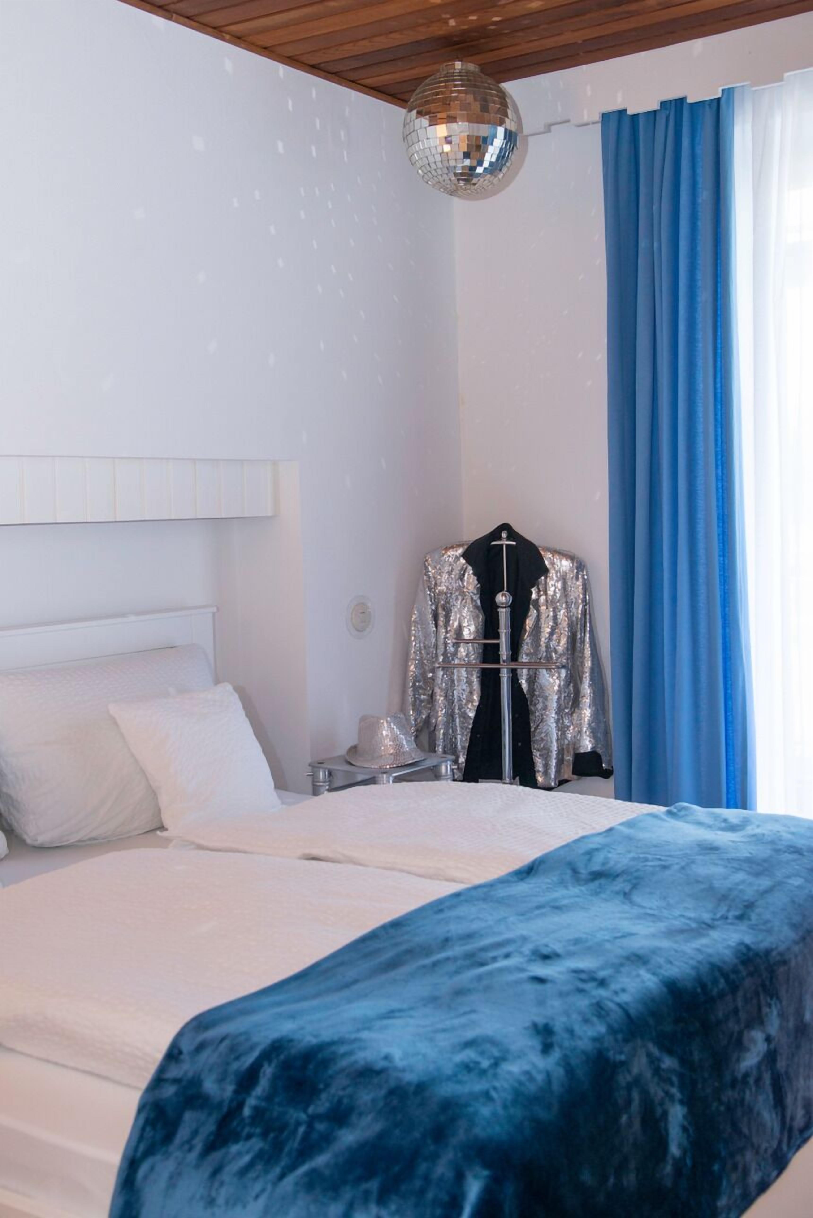 A bedroom with a bed, a blue velvet bedspread, a disco ball, a metallic jacket on a hanger, and blue curtains.