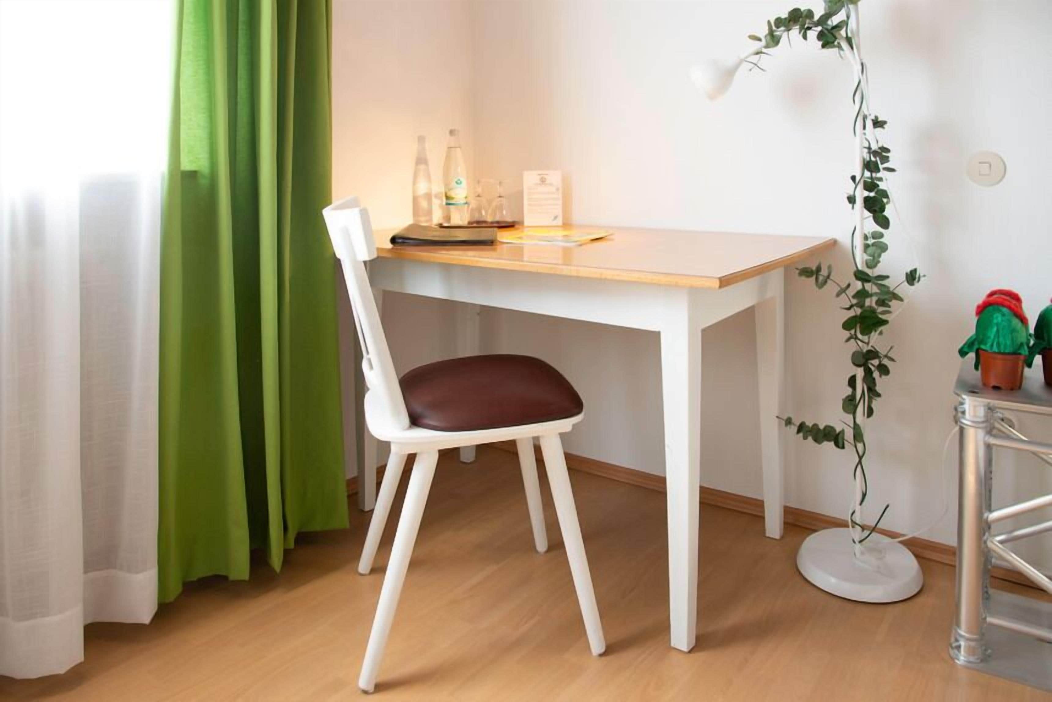 Une petite pièce propre, avec un bureau en bois, une chaise blanche, une plante en pot et un rideau vert.