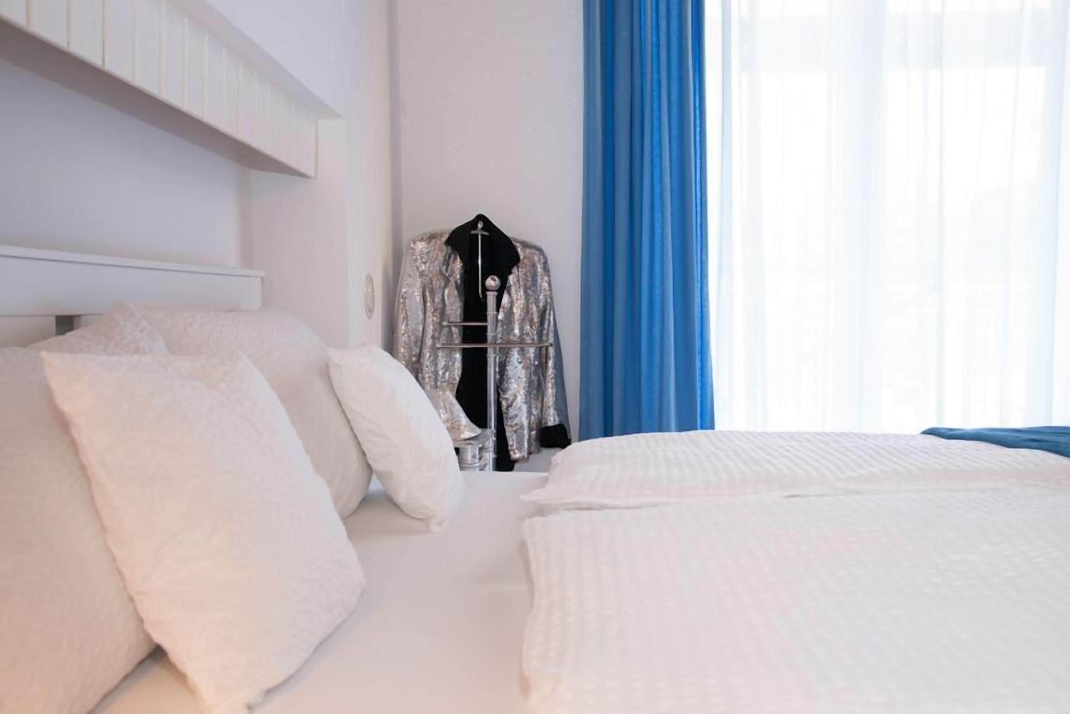 A hotel room with a bed, white bedding, a hanging coat rack, and a window with blue curtains.