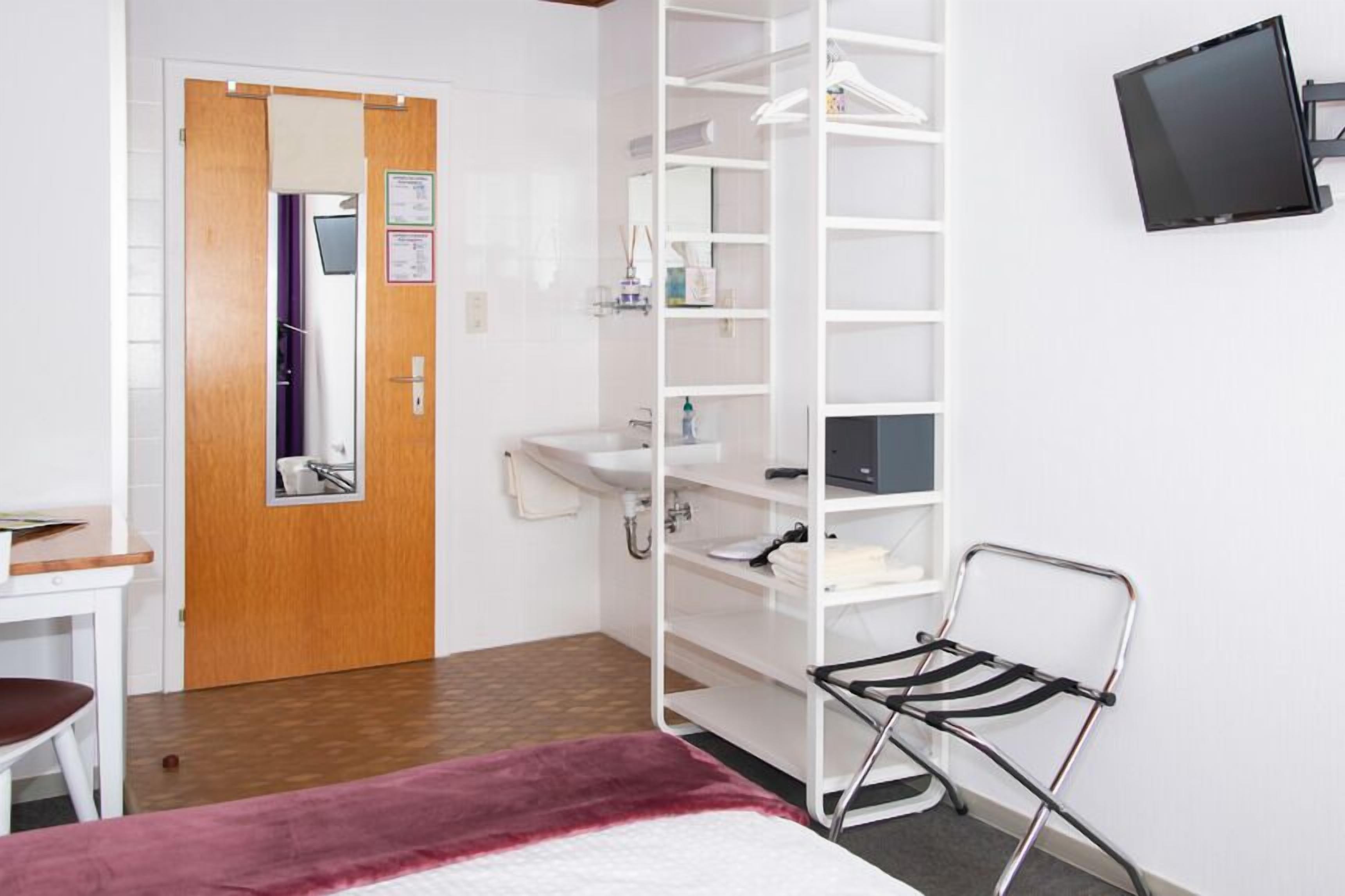 Habitación de hotel con puerta de madera, armario blanco con estantes, lavamanos pequeño, una silla y televisor de pantalla plana montado en la pared.