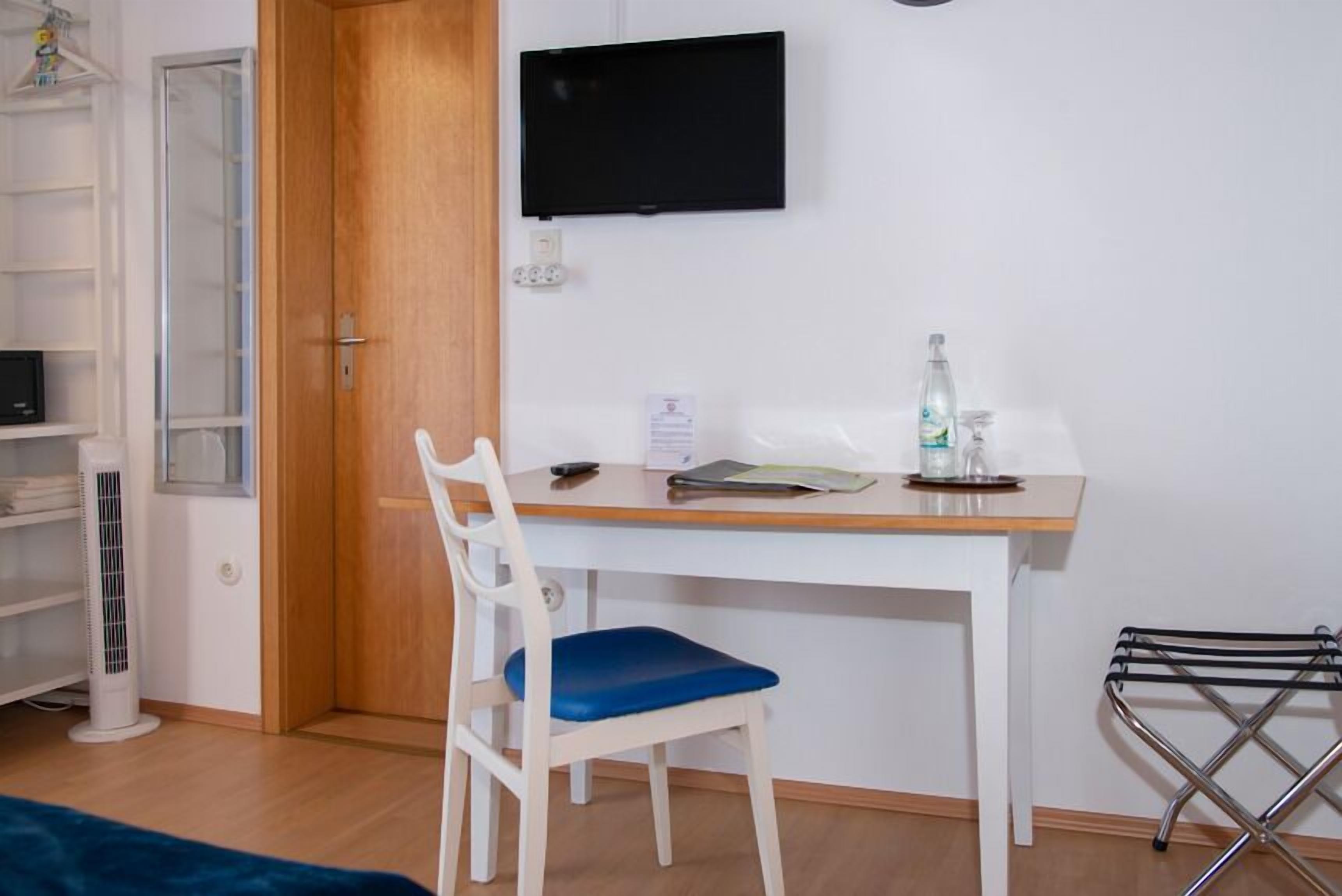A room with a white desk, a chair, a TV mounted on the wall, a radiator, a shelf with items, and a door.