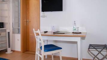 A room with a white desk, a chair, a TV mounted on the wall, a radiator, a shelf with items, and a door.