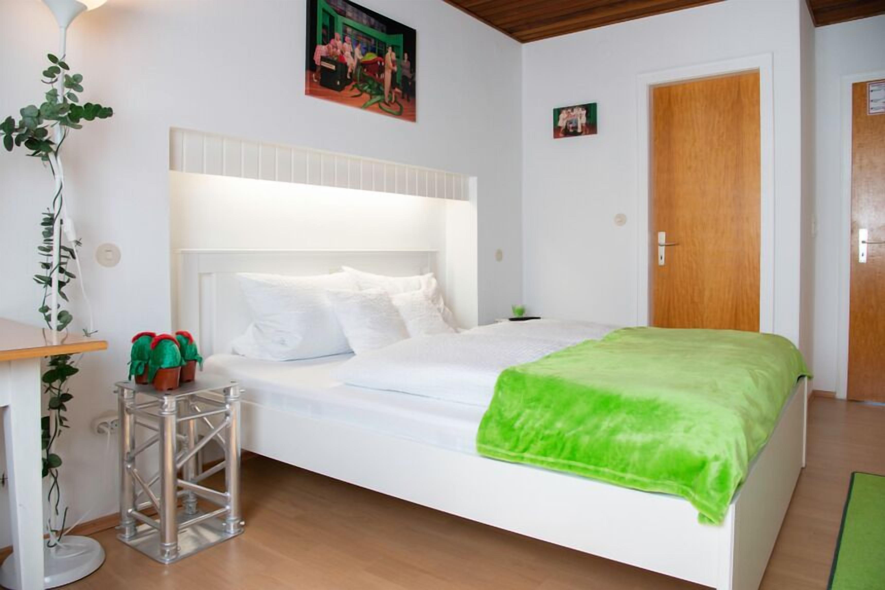 Een slaapkamer met een wit bed, een groene deken, een metalen krukje met rode objecten, een potplant en een houten deur.