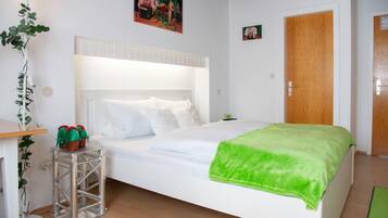 Een slaapkamer met een wit bed, een groene deken, een metalen krukje met rode objecten, een potplant en een houten deur.