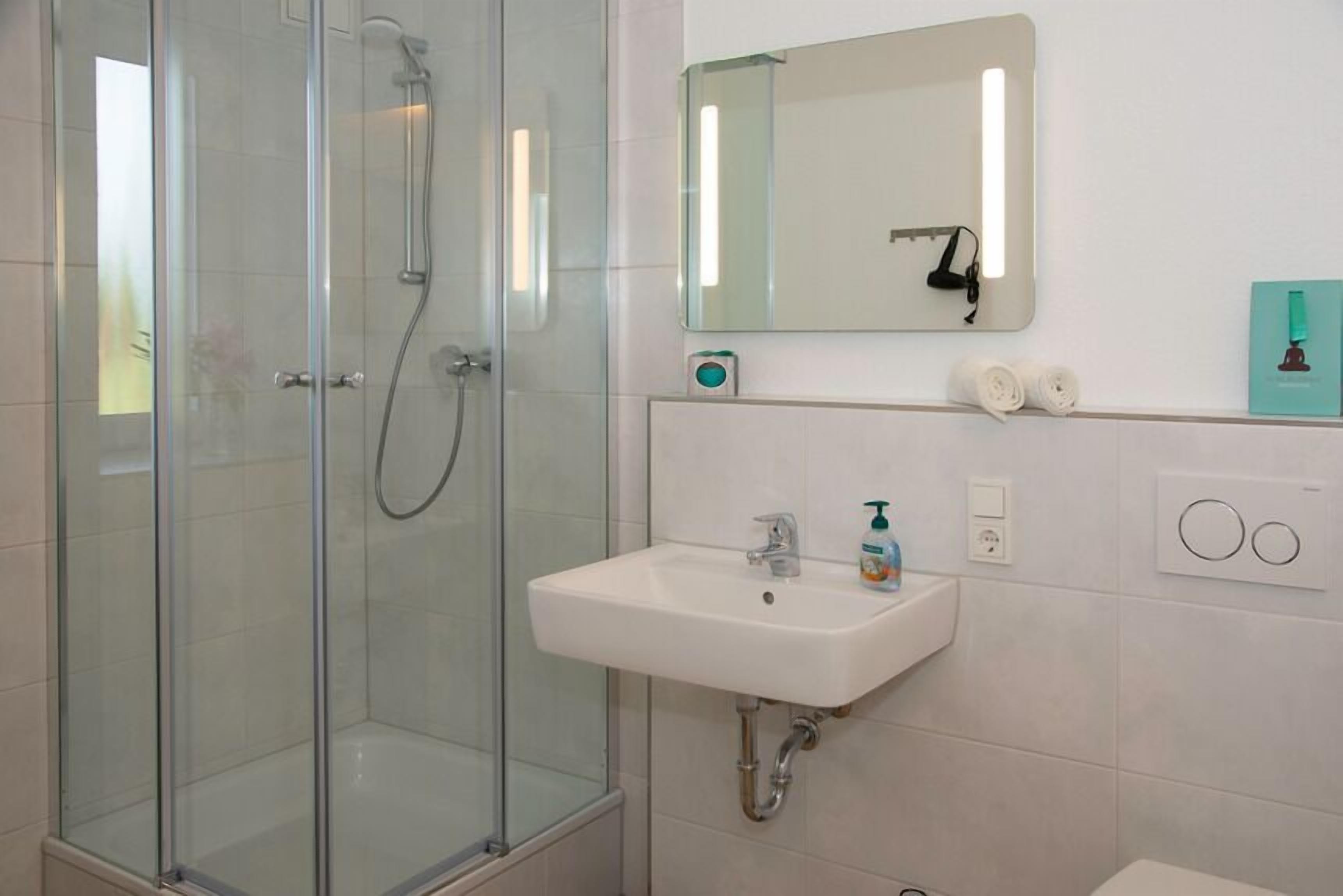 Un baño moderno con ducha envuelta en vidrio, un lavamanos blanco con espejo arriba y un inodoro a la derecha.