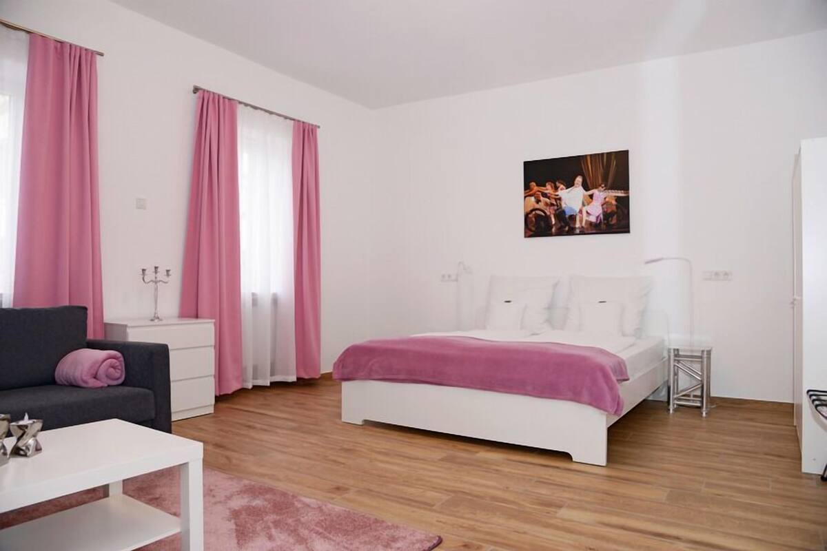 A modern bedroom with a white bed, a grey sofa, and pink curtains.