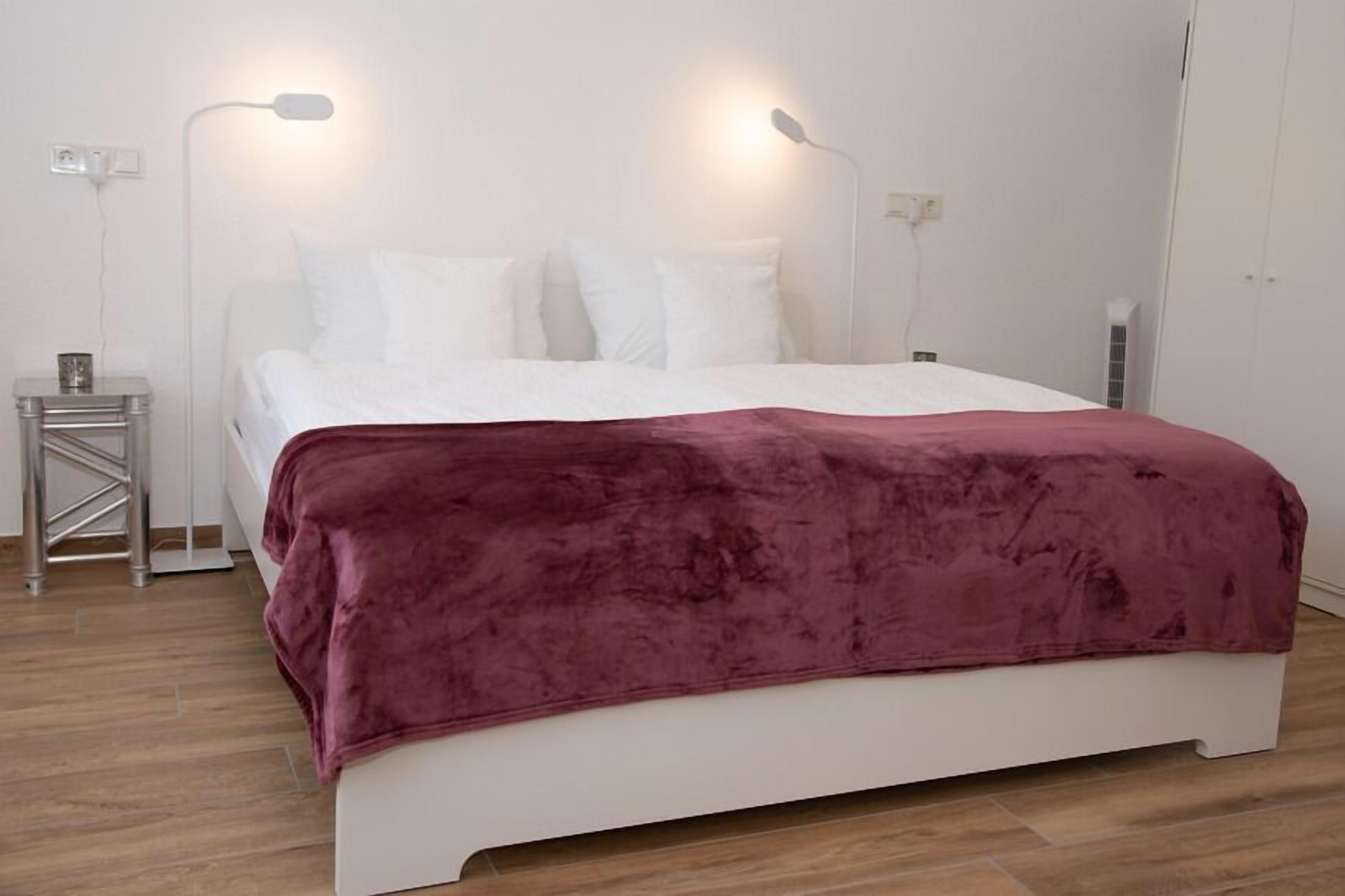 Una cama bien hecha con cabecera blanca y manta color burdeos, dos lámparas de noche y una mesita de noche con una taza.