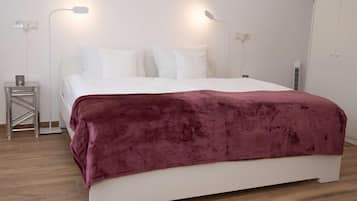 Una cama bien hecha con cabecera blanca y manta color burdeos, dos lámparas de noche y una mesita de noche con una taza.
