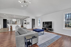 A spacious living room with a grey sofa, a blue ottoman, a TV, and a kitchen in the background. - Chamblee Retreat 3BR Home w Spacious Porch (Chamblee)