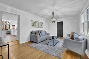 A spacious living room with a grey sofa, a coffee table, and a chandelier. There is a kitchen visible in the background. - Chamblee Retreat 3BR Home w Spacious Porch (Chamblee)