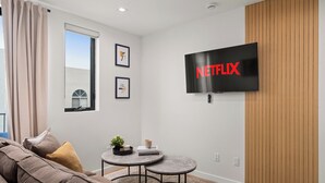 Ein modernes Wohnzimmer mit einem großen Flachbildfernseher, der das Netflix-Logo anzeigt, einem braunen Sofa, einem kleinen runden Tisch mit einer Pflanze und gerahmten Kunstwerken an der Wand.