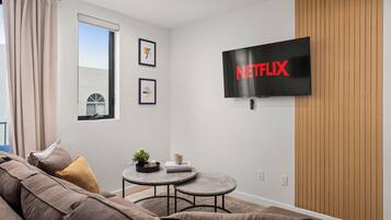 Un salon moderne avec un grand téléviseur à écran plat affichant le logo de Netflix, un canapé marron, une petite table ronde avec une plante et des œuvres d’art encadrées accrochées au mur.