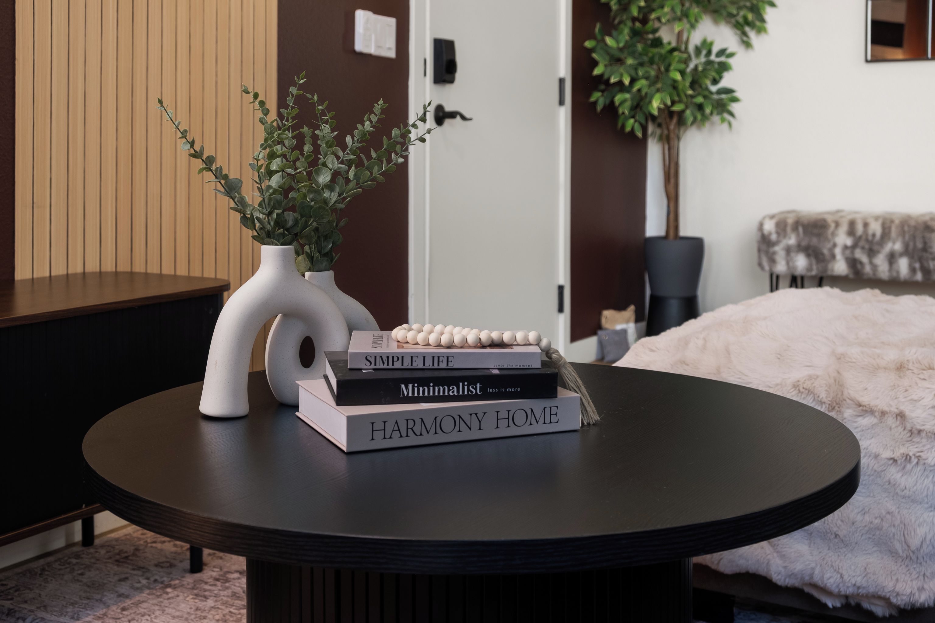 A round table with a white vase and books, a door, and a plant in the background.