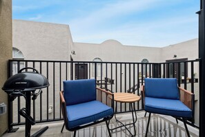 A patio with two blue cushioned chairs and a small table, a grill, and a view of a building with arches. - King Bed, EV Charger, Sleeps 8, Balcony, Laundry (San Diego)