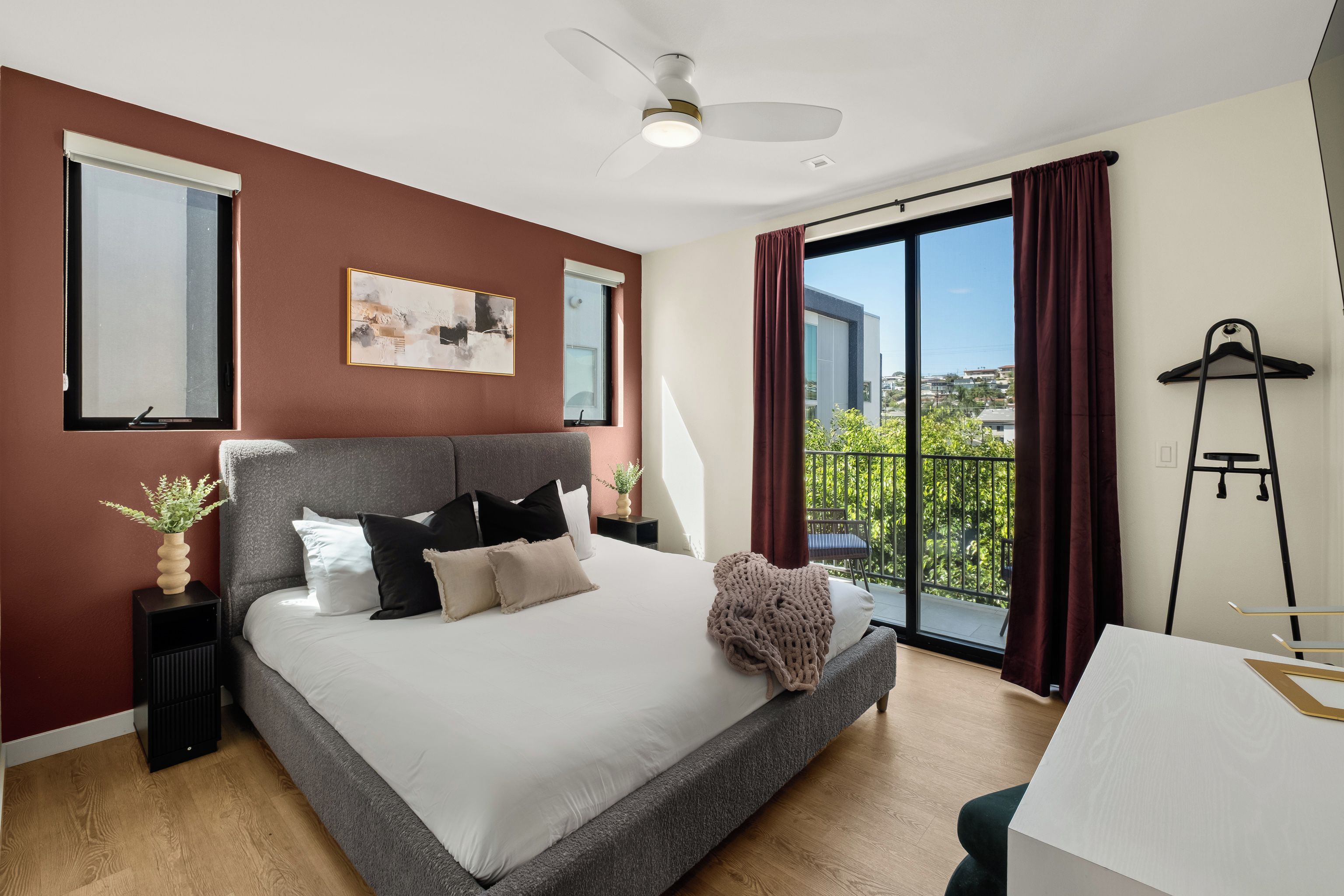A modern bedroom with a large bed, a ceiling fan, a window with curtains, and a balcony with a view.