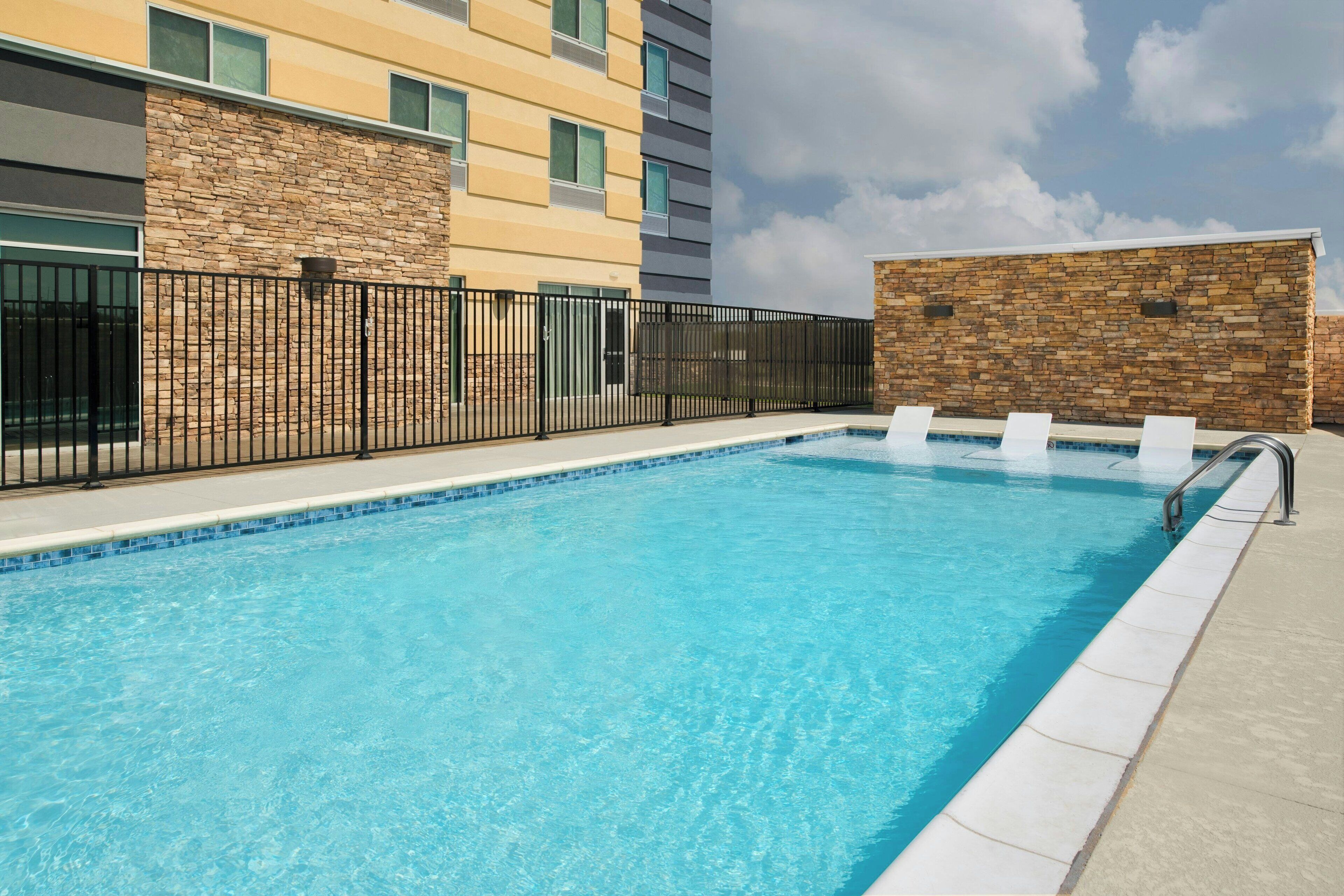 A rectangular swimming pool with clear blue water, surrounded by a black metal fence and a stone wall.