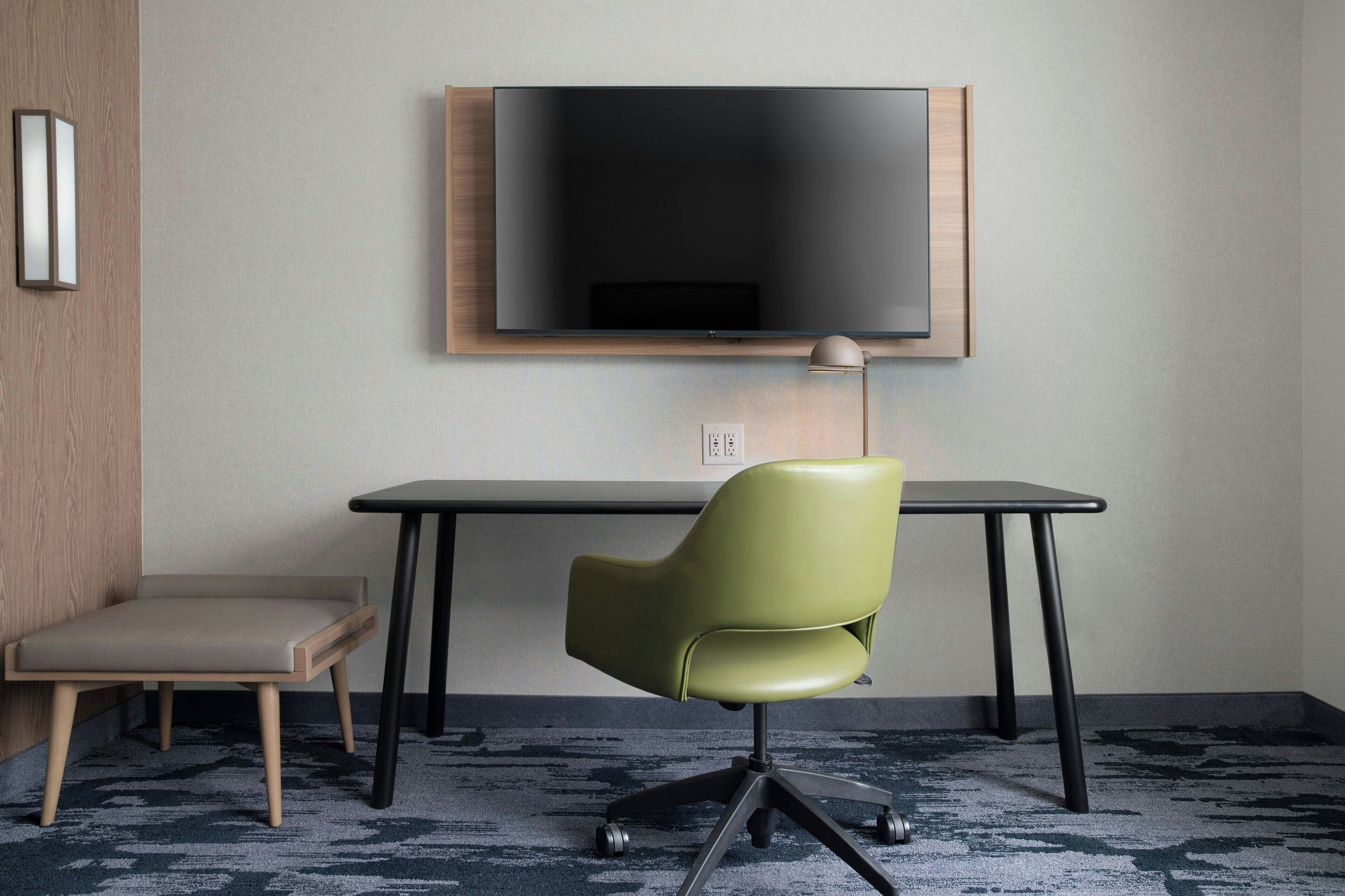 Una habitación moderna con una televisión de pantalla plana montada en la pared, una silla de oficina verde, un pequeño banco de madera y un escritorio con una lámpara.