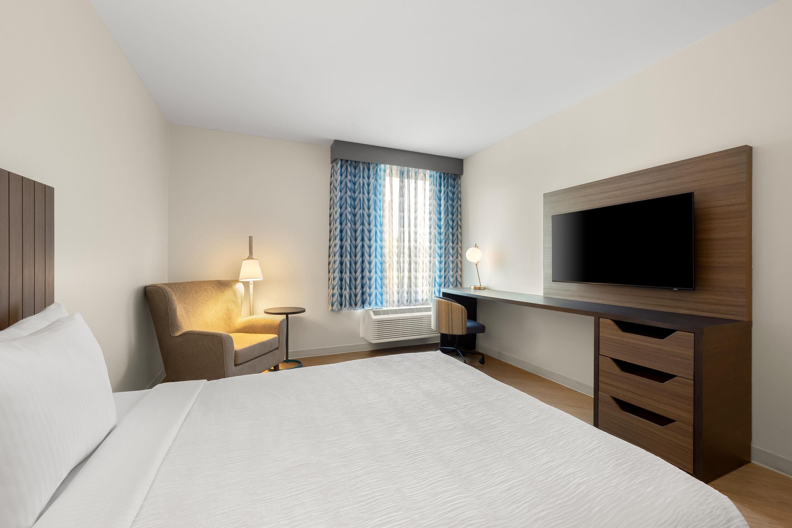 Una habitación de hotel con cama, escritorio, televisor de pantalla plana, silla, lámpara y ventana con cortinas.