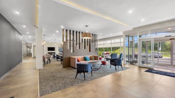 A modern hotel lobby with a seating area, a fireplace, and a large window offering a view of the outdoors.