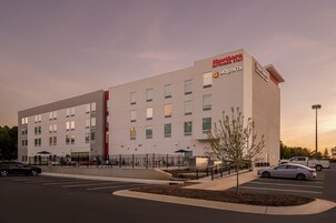 A modern hotel building with multiple floors, a parking lot, and a landscaped area with a small tree.
