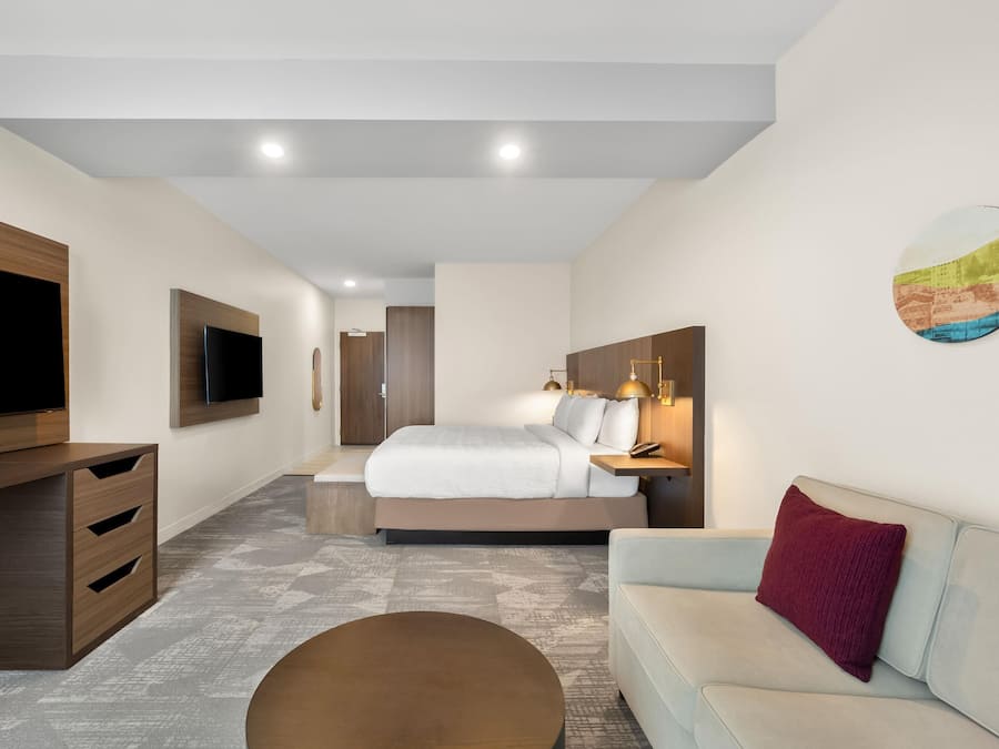 A modern hotel room with a flat-screen TV, a bed with a headboard, a sofa, a coffee table, and a painting on the wall.