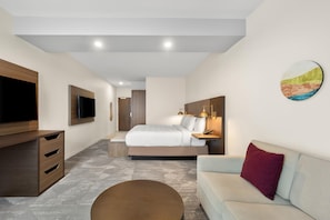 A modern hotel room with a flat-screen TV, a bed with a headboard, a sofa, a coffee table, and a painting on the wall. - La Quinta Inn & Suites by Wyndham Mebane (Mebane)
