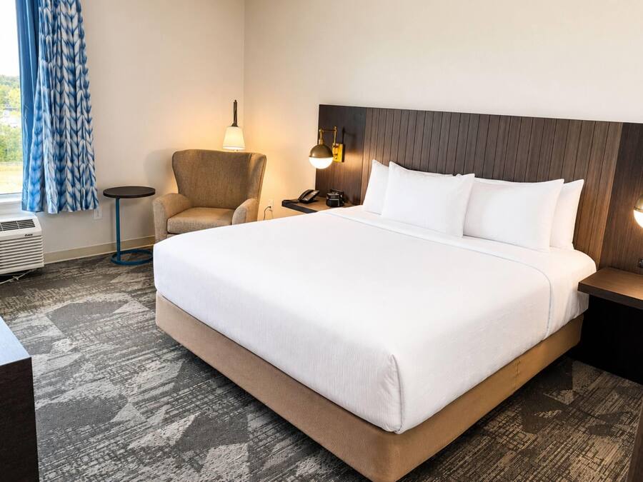 Une chambre d’hôtel avec un grand lit, une chaise, un bureau, une lampe, un téléphone et une fenêtre avec des rideaux.