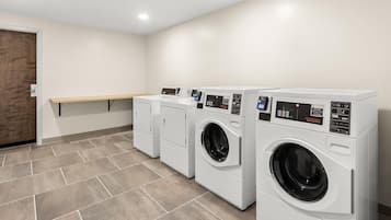 A laundry room with three white commercial washers and dryers, a wooden bench, and a door.
