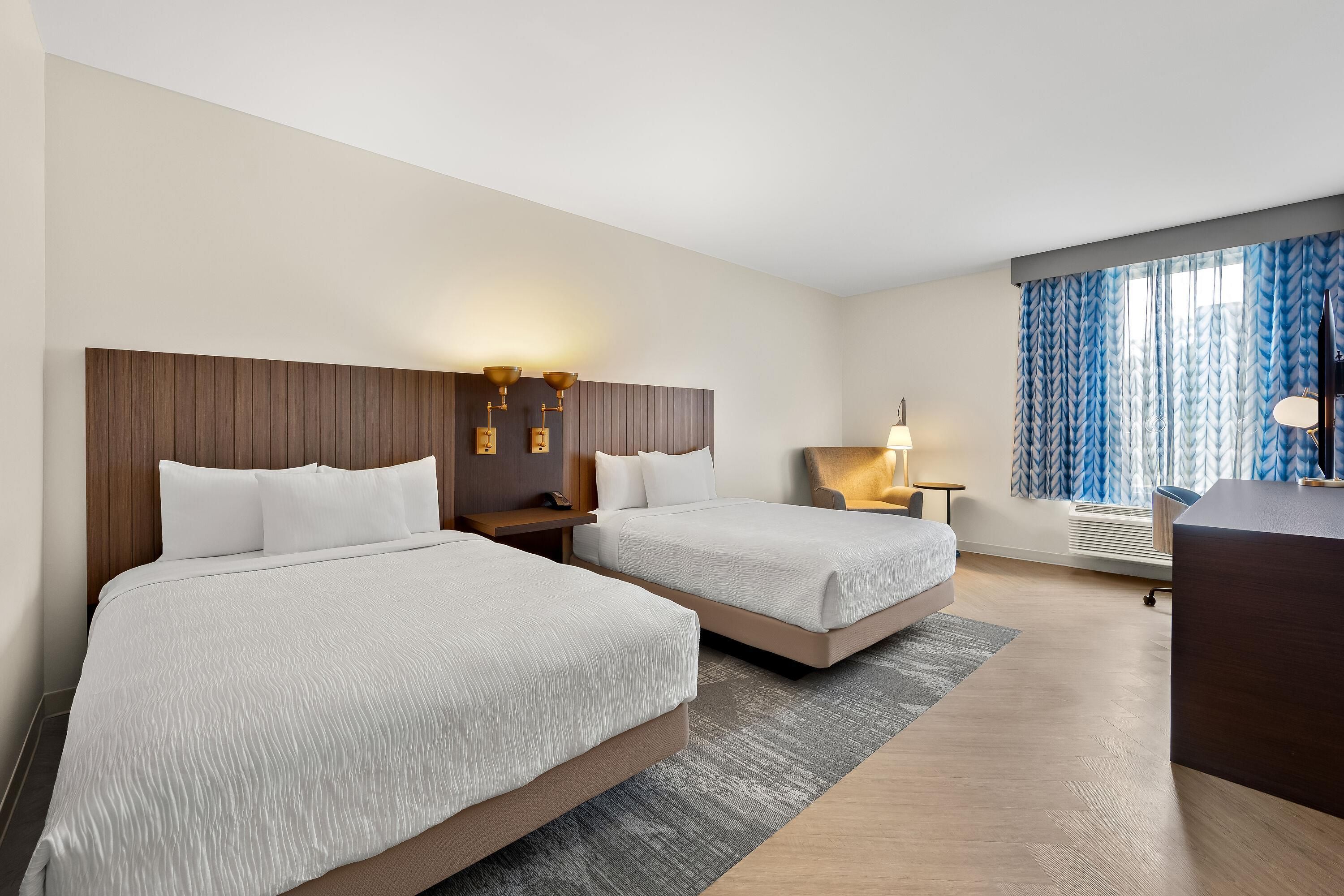 A hotel room with two beds, a desk, a chair, a lamp, and a window with curtains.