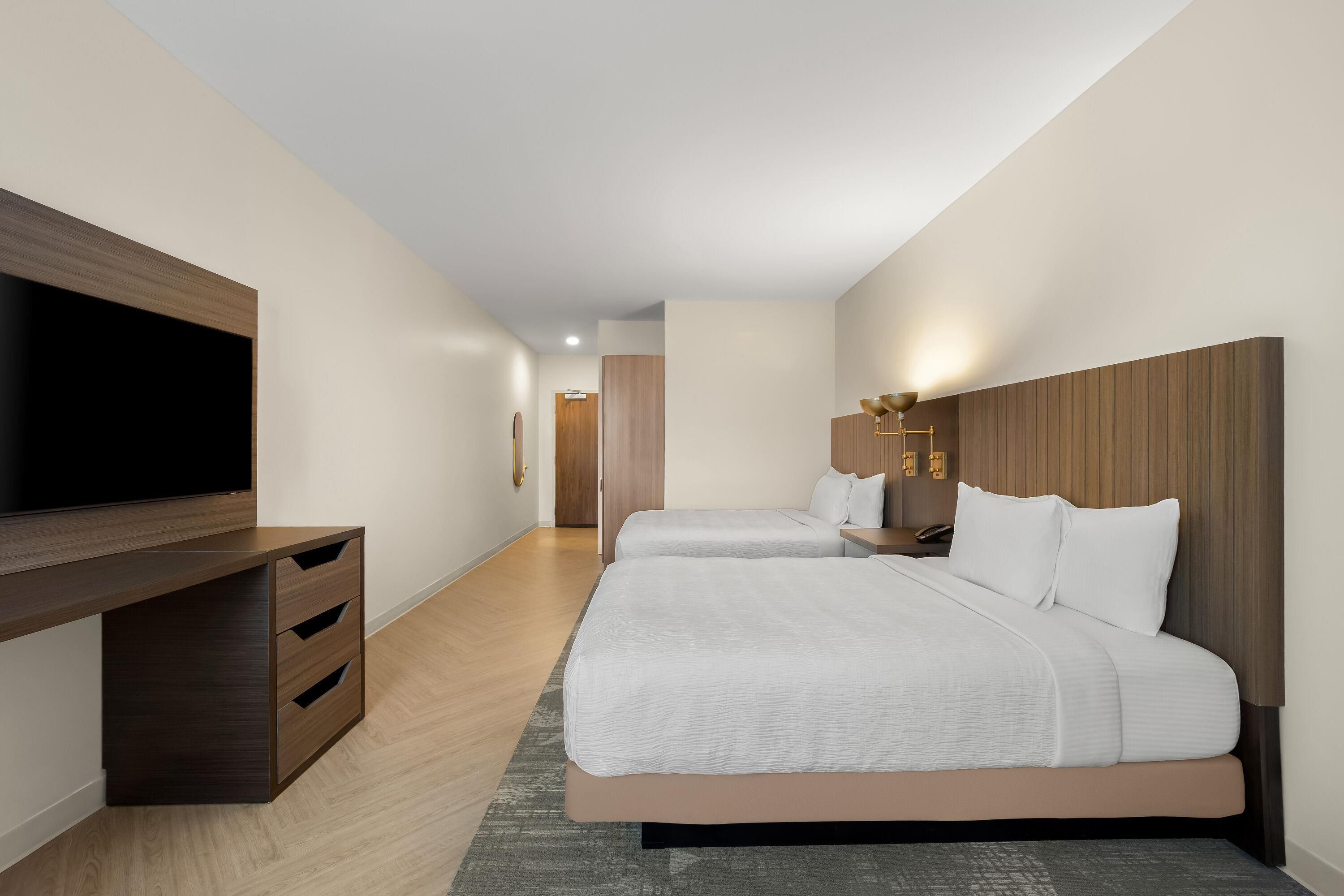 Quarto de hotel com uma televisão de tela grande, duas camas com roupa de cama branca, uma mesinha de cabeceira de madeira e uma luminária fixada na parede.