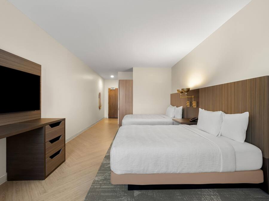 Quarto de hotel com uma televisão de tela grande, duas camas com roupa de cama branca, uma mesinha de cabeceira de madeira e uma luminária fixada na parede.
