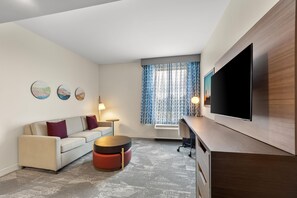A modern living room with a sofa, a coffee table, a flat-screen TV, and a patterned window. - La Quinta Inn & Suites by Wyndham Mebane (Mebane)