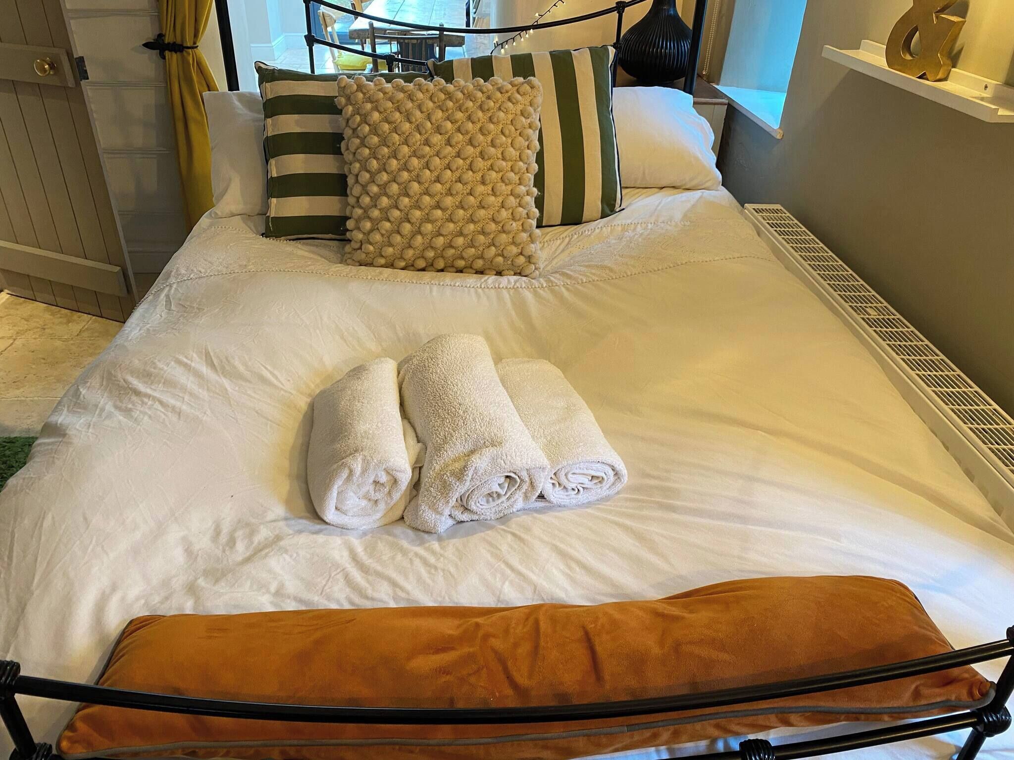 A neatly made bed with white linens, two rolled towels, and a brown throw pillow.