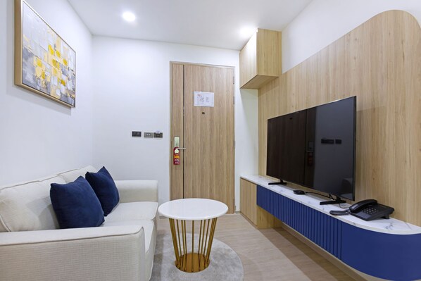 A modern hotel room with a white sofa, a round table, a flat-screen TV, and a reception desk.
