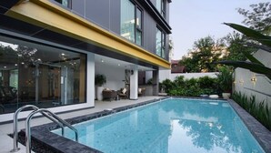 Un espace piscine moderne avec une piscine à eau claire, une poutre jaune au-dessus et un patio vitré aménagé avec du mobilier.