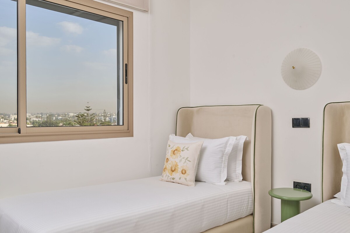 A room with two beds, a window, a green side table, and a wall-mounted light fixture.