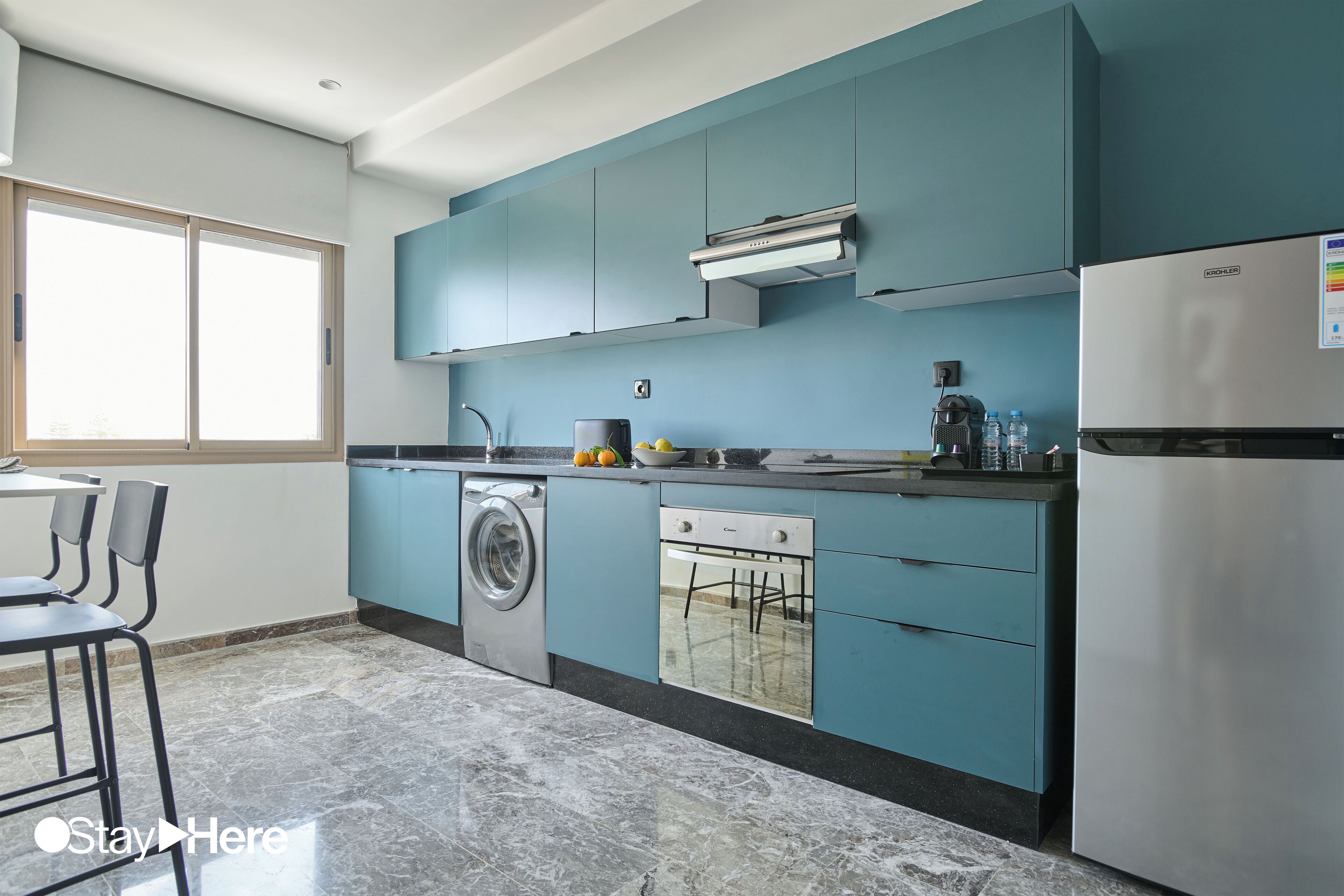 Une cuisine moderne avec des meubles de couleur turquoise, un réfrigérateur en acier inoxydable, un lave-linge encastré et un coin repas agrémenté de tabourets de bar.
