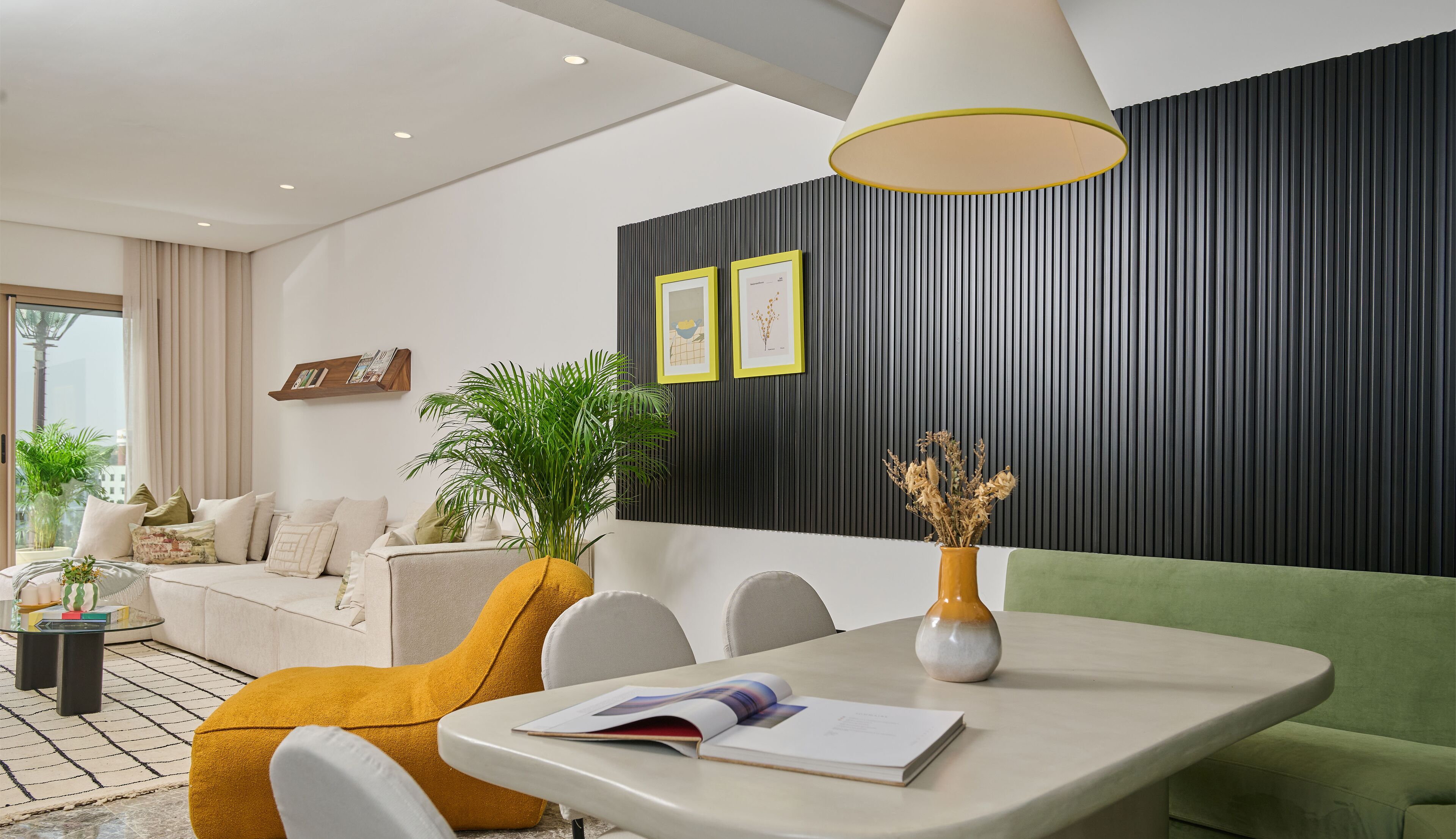 A modern living room with a sofa, a dining table with a book, a potted plant, and a wall with vertical panels.