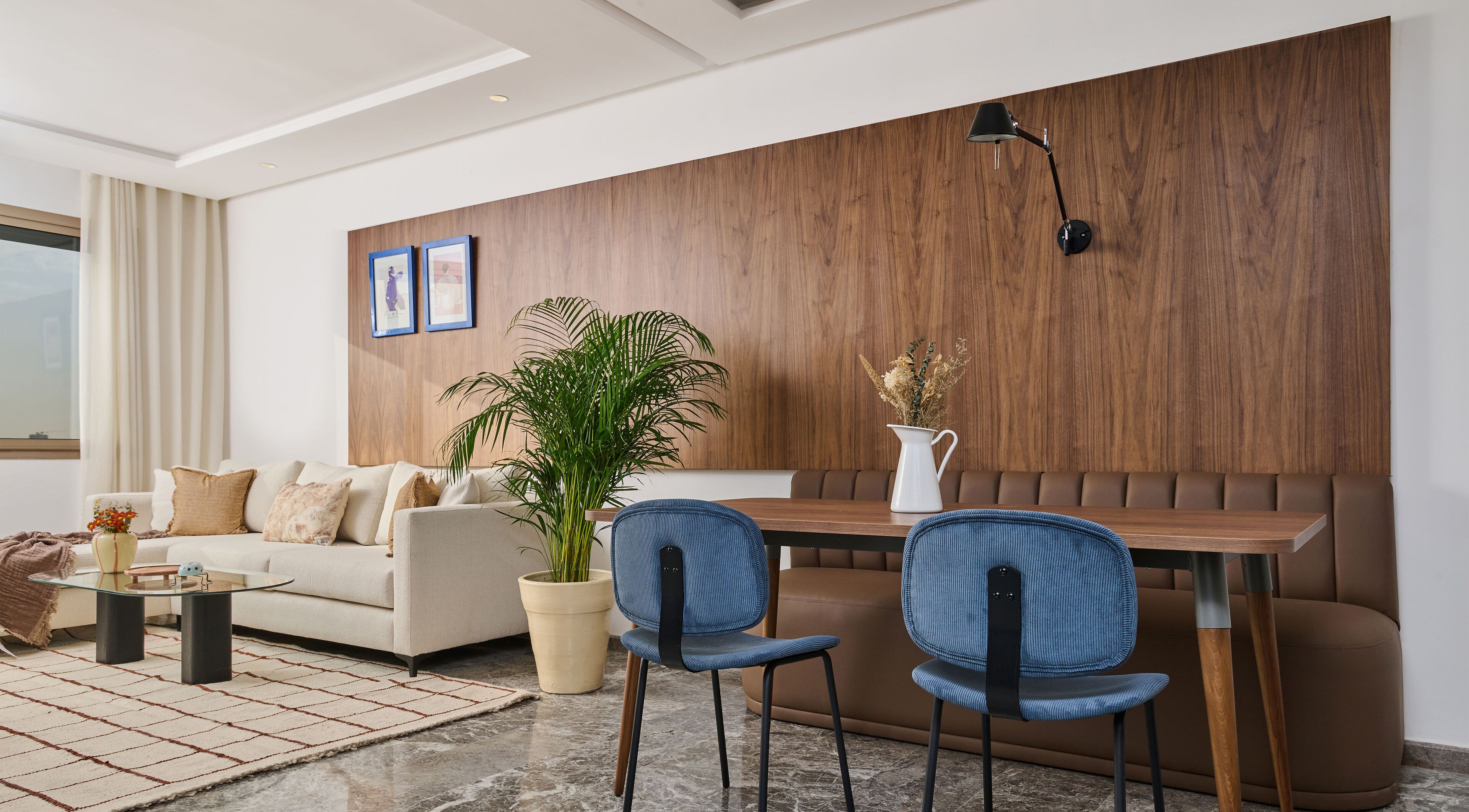 Uma sala de estar moderna com um sofá, mesa de centro e cadeiras. Há uma parede de destaque em madeira, uma planta em vaso e um vaso com flores.