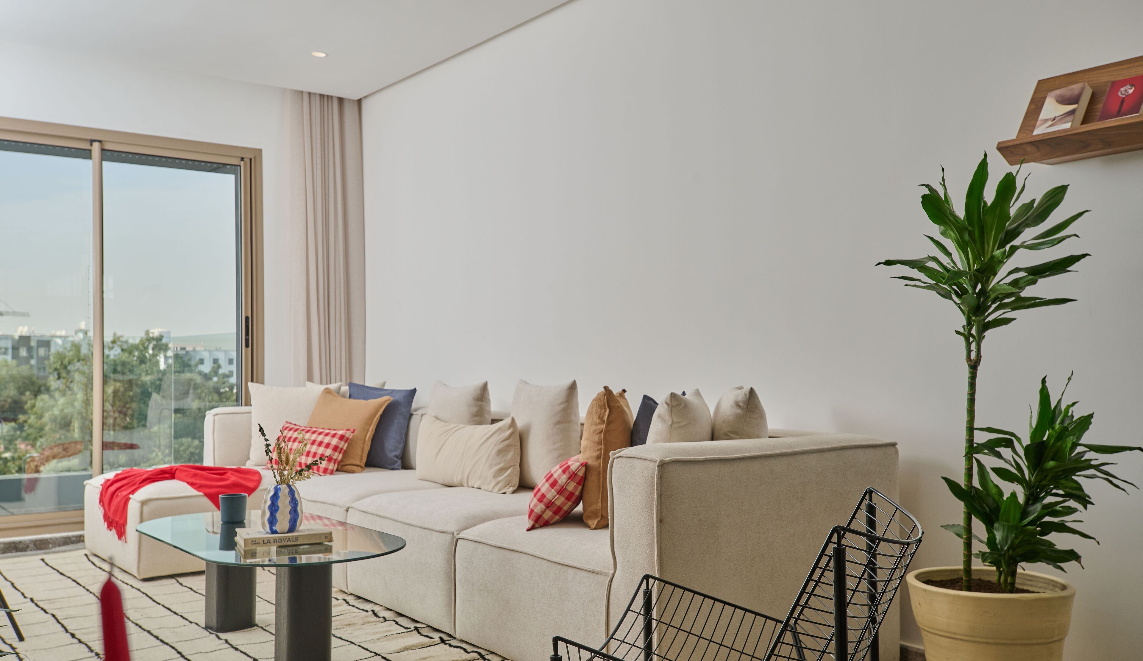A modern living room with a sofa, a glass coffee table, a potted plant, and a view of the city through large windows.