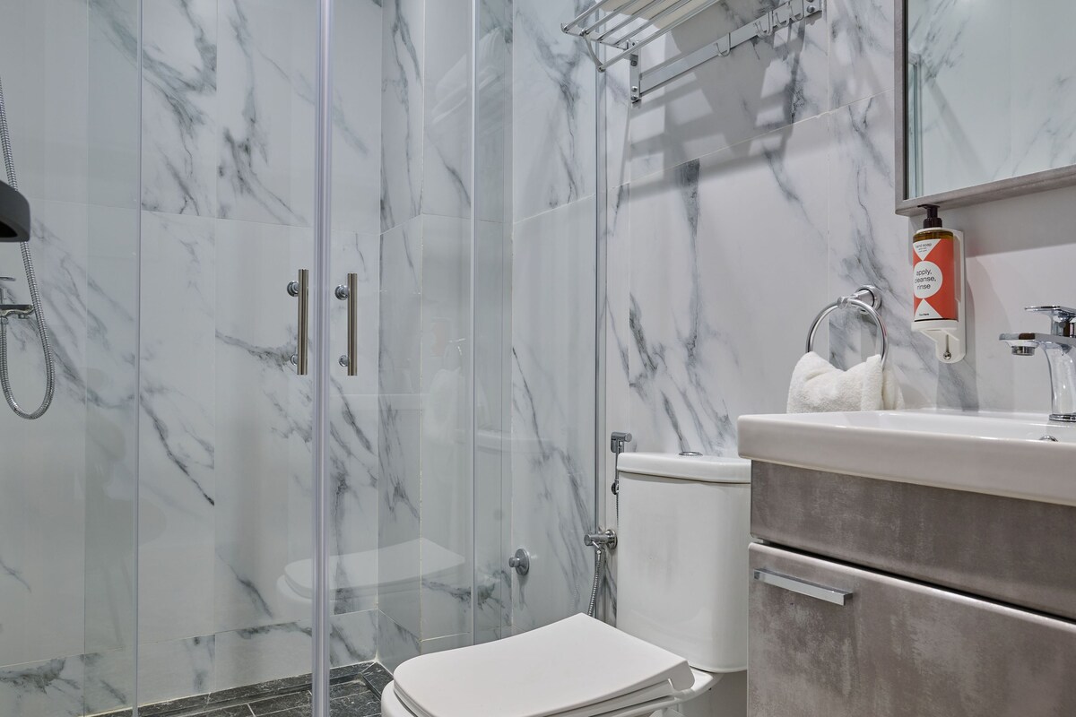 Un baño con ducha envuelta en vidrio, inodoro blanco y lavamanos con dispensador de jabón.