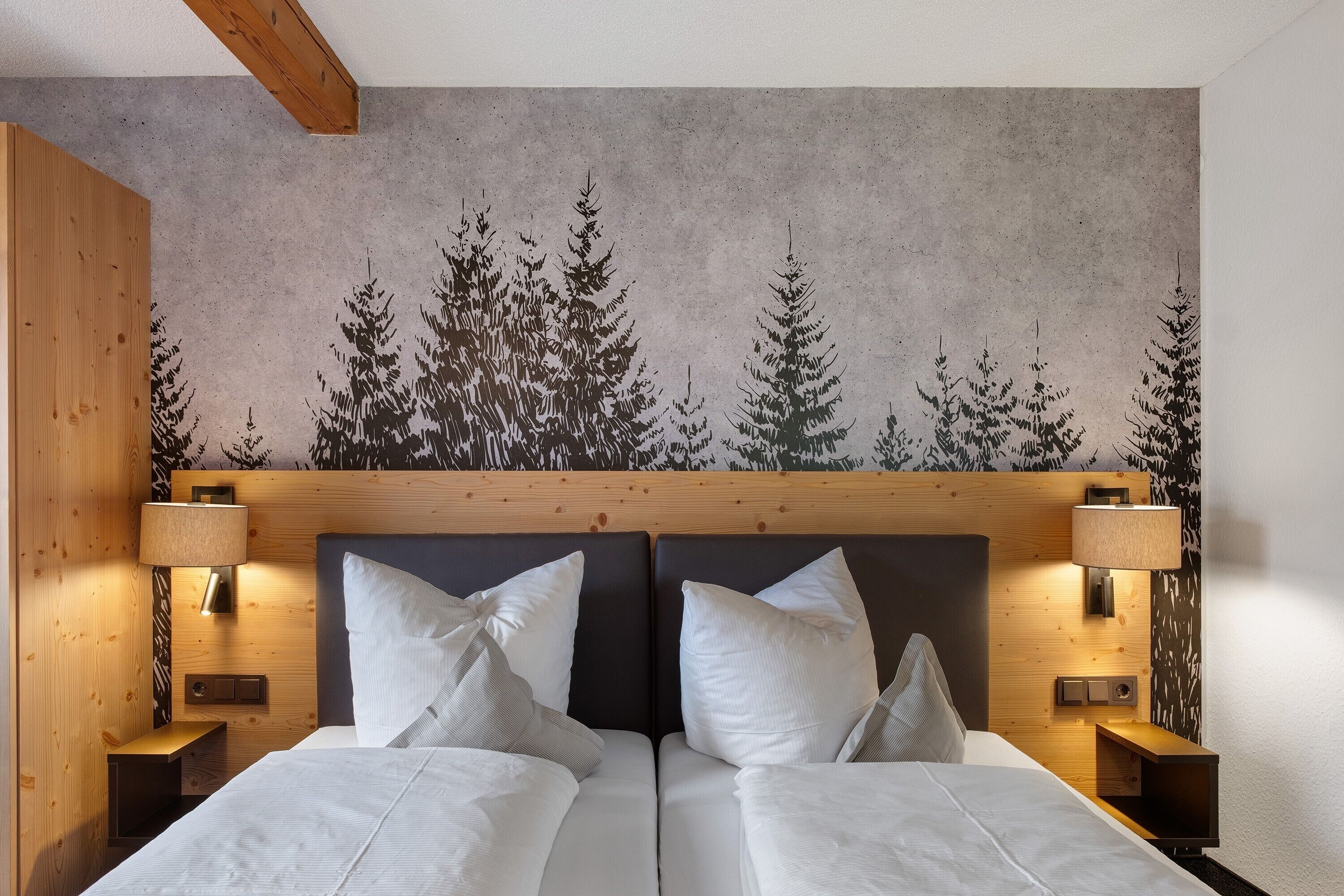 Un lit double avec du linge de lit blanc, deux tables de chevet avec des lampes et une tête de lit ornée d’une fresque représentant une forêt.