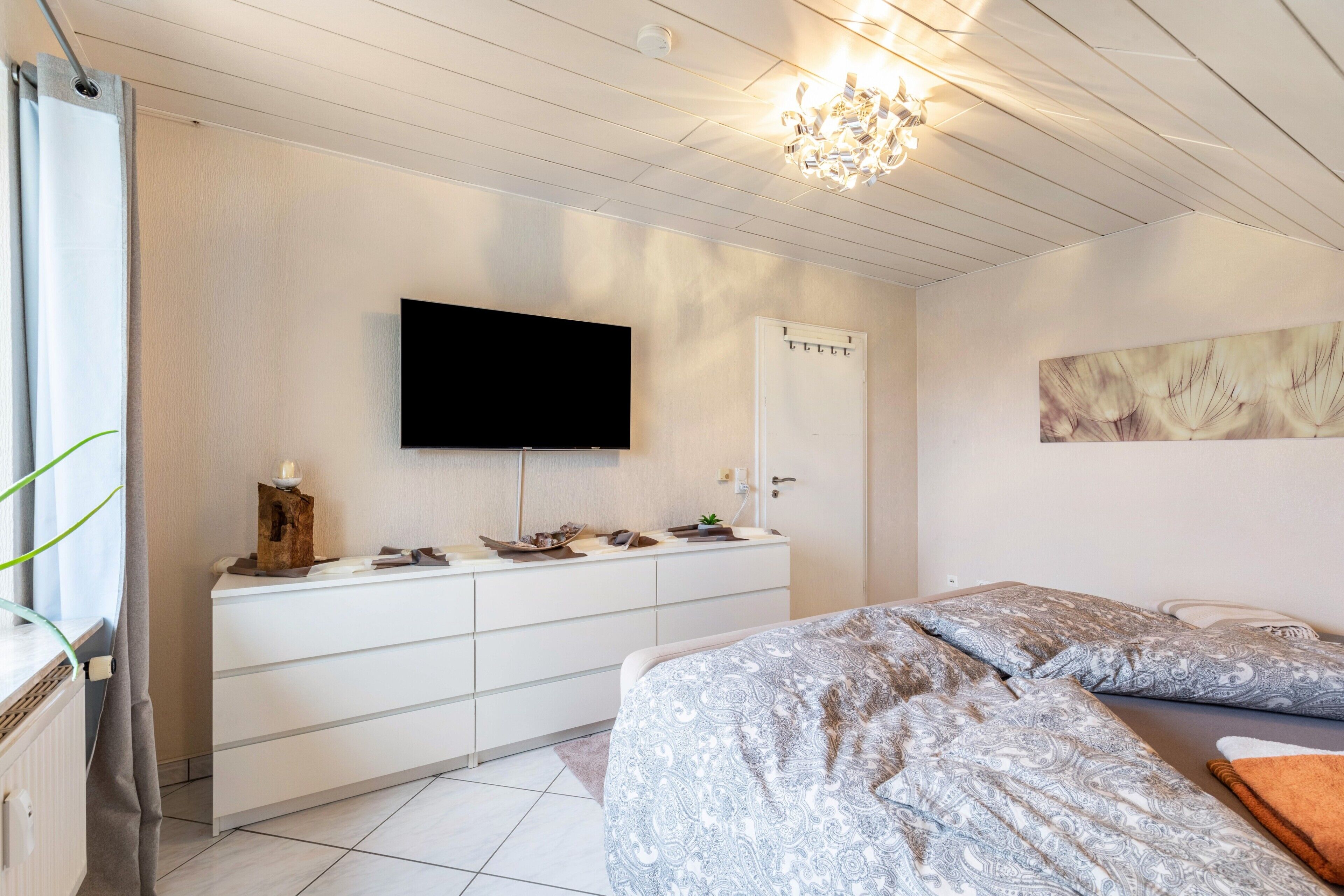 Un dormitorio con cama, televisor montado en la pared, escritorio con cajones y ventana con cortinas.