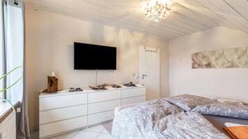 Un dormitorio con cama, televisor montado en la pared, escritorio con cajones y ventana con cortinas.