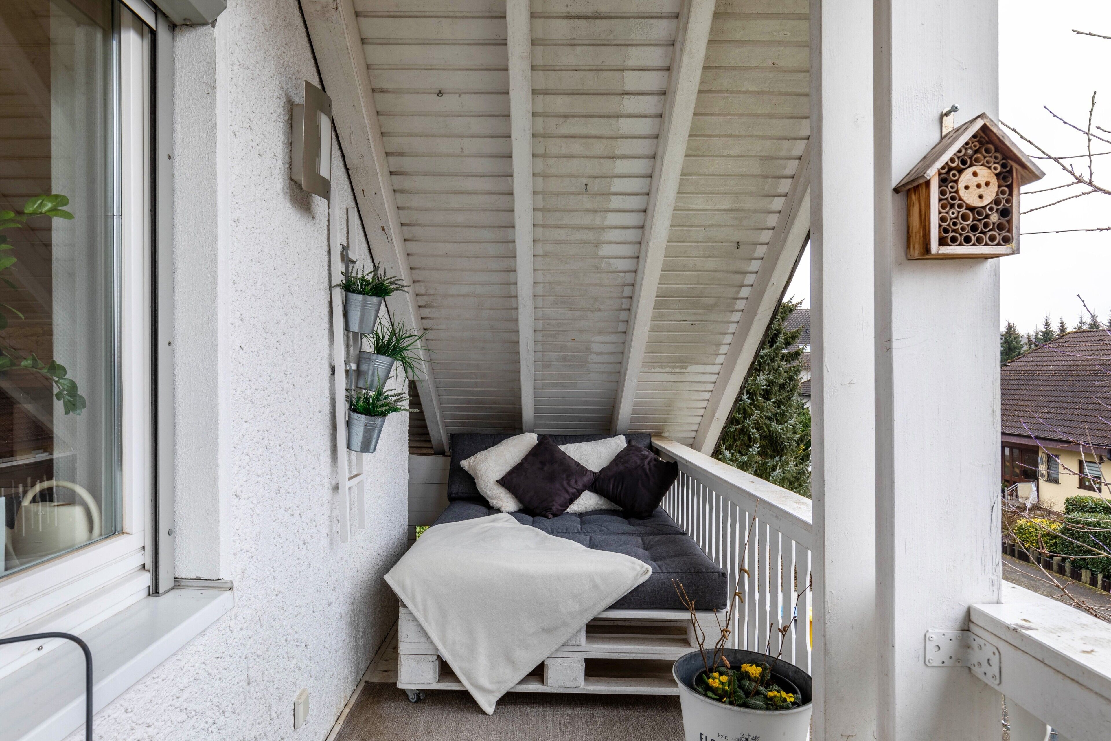 A balcony with a cushioned seating area, a hanging plant, and a birdhouse.