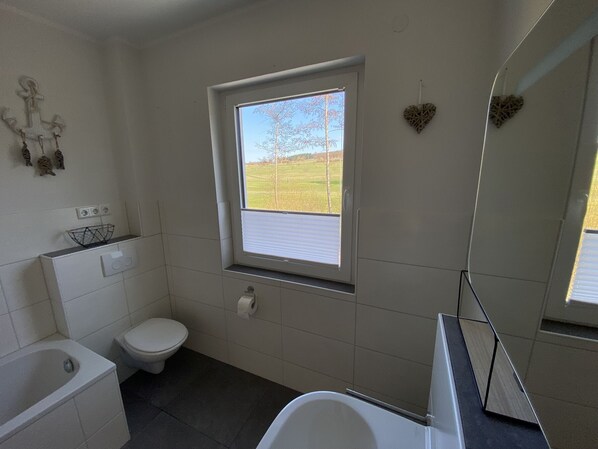 A bathroom with a toilet, a bathtub, a window with a view of a field, and a mirror.