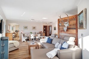 Un salon moderne avec un canapé, une table basse et une tête de cerf fixée au mur. On y trouve également un coin repas avec des chaises et une table, et une cuisine à l’arrière-plan.